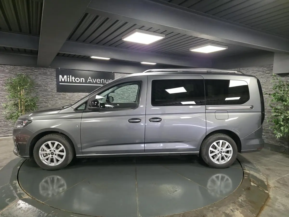 Vue de profil côté gauche d’un Ford Tourneo Connect gris métallisé avec barres de toit argentées en intérieur showroom.