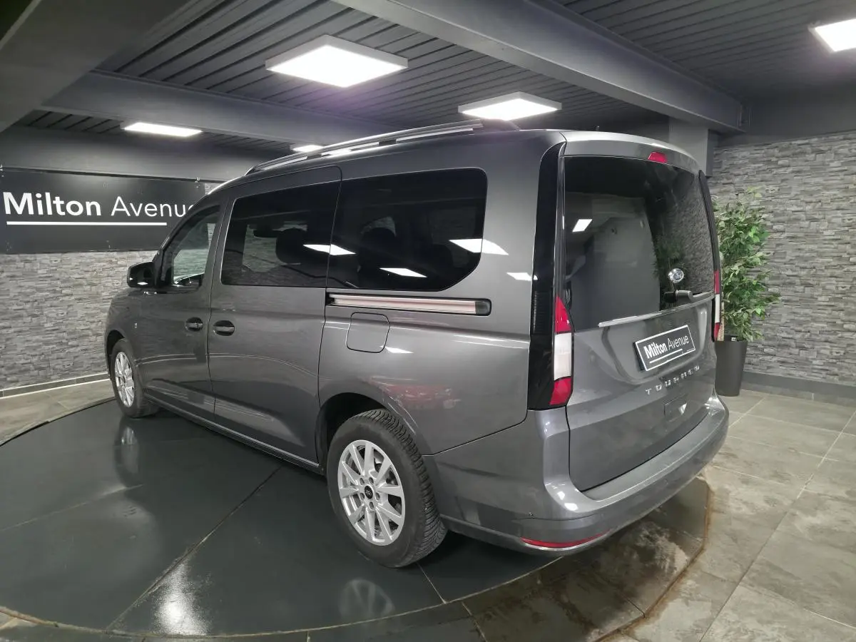 Vue 3/4 arrière droit du Ford Transit Connect Tourneo gris métallisé avec barres de toit argentées en intérieur showroom.