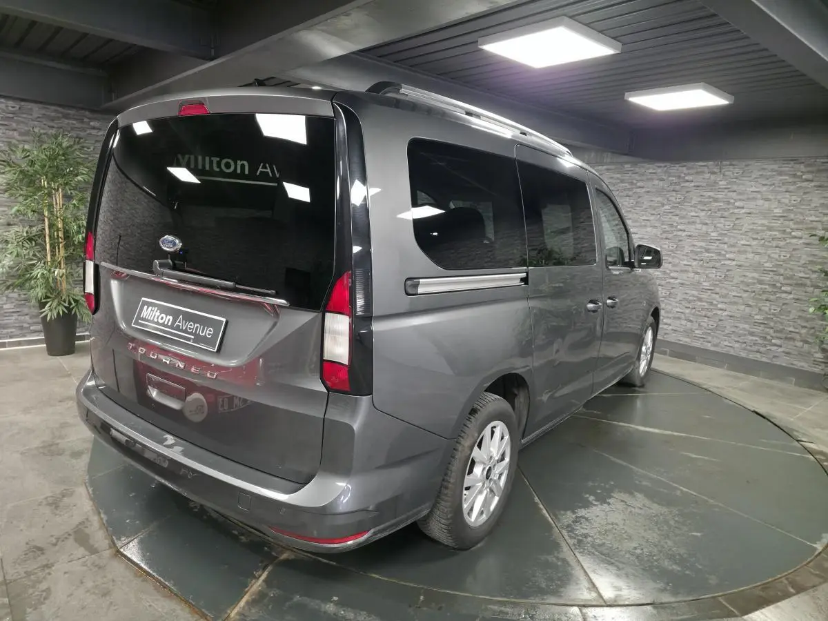 Vue 3/4 arrière droite du Ford Tourneo Connect gris métallisé avec vitres teintées et barres de toit argentées.