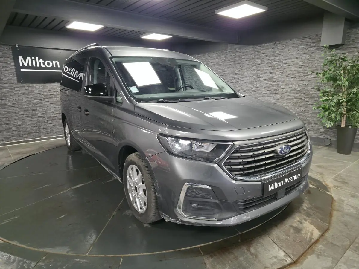 Vue 3/4 avant droit du Ford Transit Connect gris métallisé avec calandre noire et barres de toit argentées en showroom.