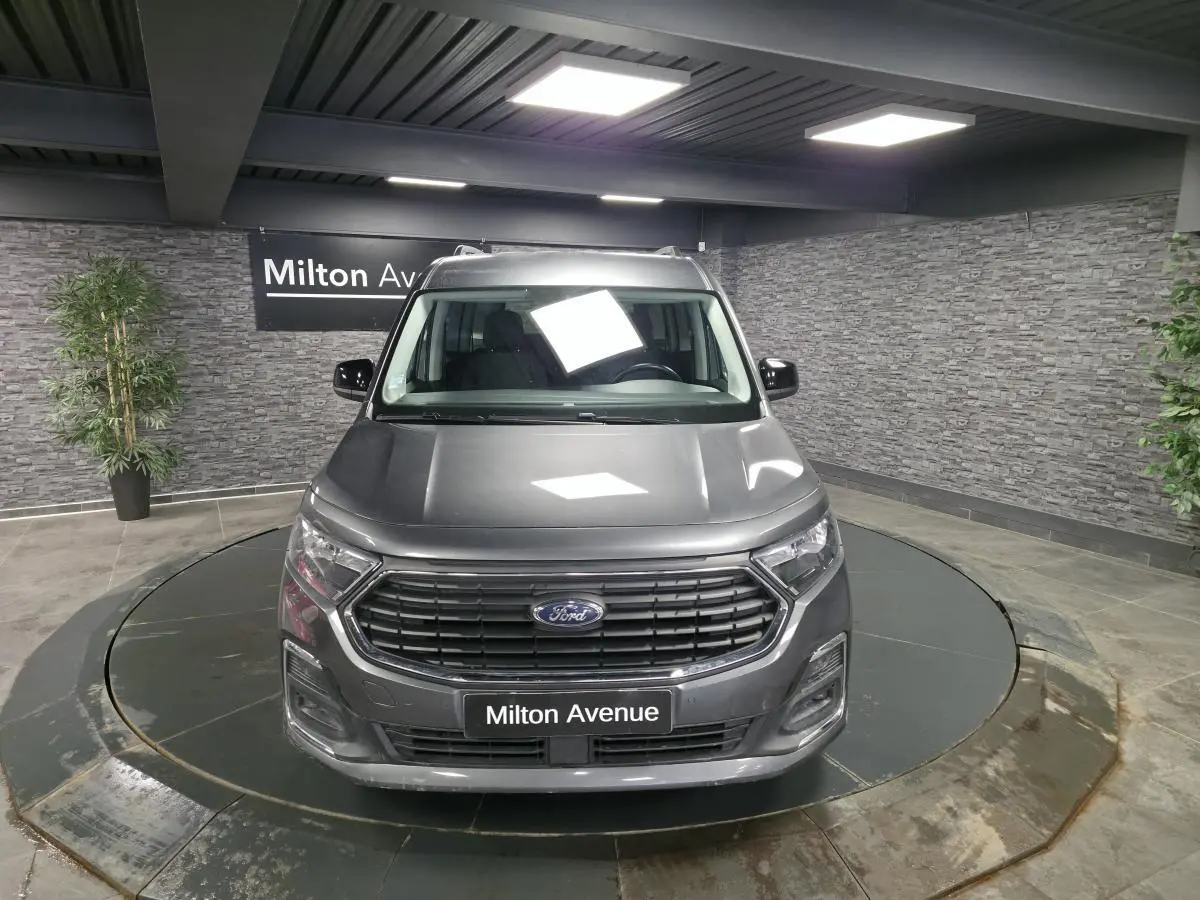 Vue avant d'un Ford Transit Connect gris métallisé avec calandre noire et barres de toit argentées en showroom.