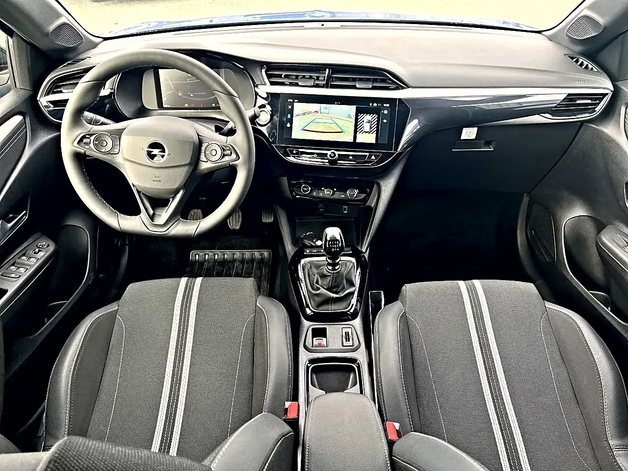 Vue intérieure avant de l’Opel Corsa 2025, tableau de bord noir avec écran tactile et sièges sport gris à bandes blanches.