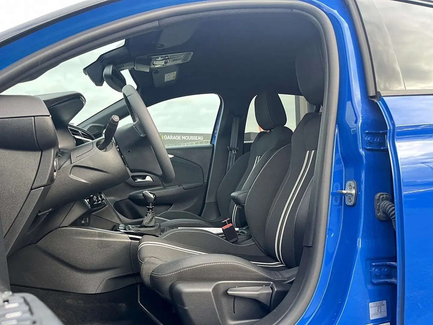 Vue intérieure côté conducteur d'une Opel Corsa bleu 2025, sièges sport noirs avec surpiqûres blanches et boîte manuelle.