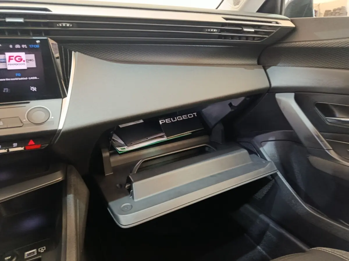 Vue intérieure côté passager de la Peugeot 308 2022, boîte à gants ouverte avec manuel visible, tableau de bord gris.