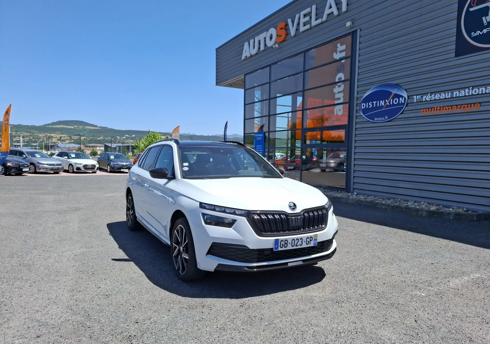 Vue 3/4 avant d'un Skoda Kamiq blanc 2021 avec jantes alliage noires devant un bâtiment commercial.
