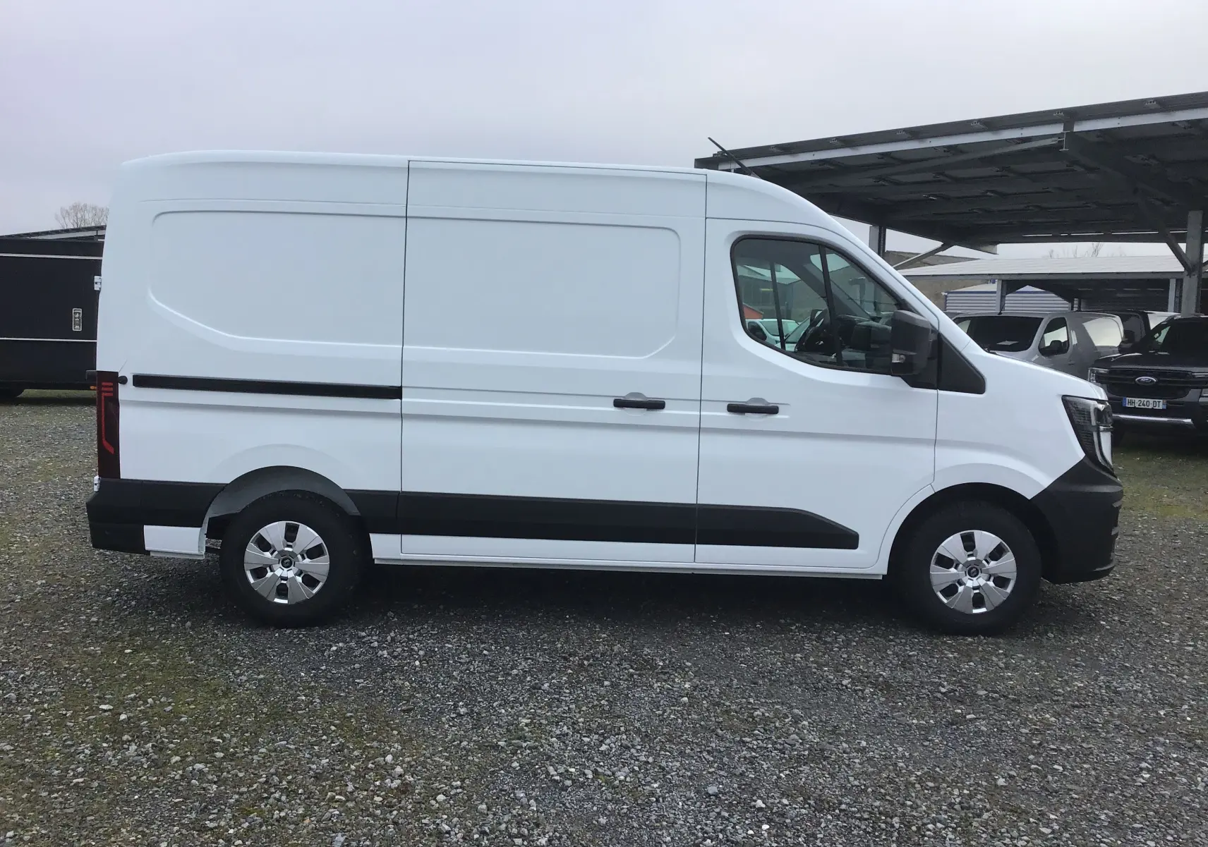 Vue latérale droite d’un Renault Master blanc L2H2 avec bandeaux noirs et jantes argentées sur un sol gravillonné.