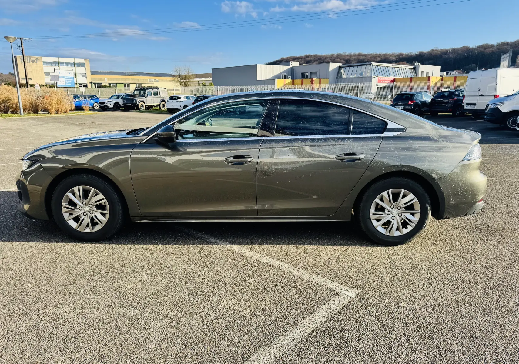 Profil côté gauche d'une Peugeot 508 gris amazonite 2019 garée en extérieur sous un ciel bleu.