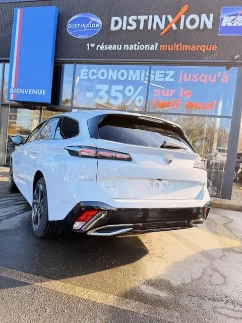 Vue 3/4 arrière droite d'une Peugeot 308 Hybrid 145 e-DCS6 Allure blanche avec feux LED et jantes noires devant un concessionnaire multimarque.