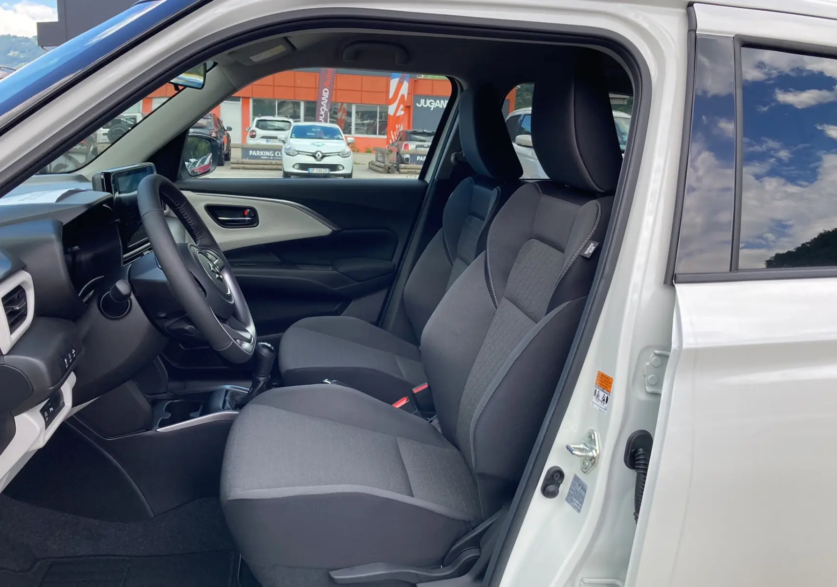 Vue intérieure côté conducteur de la Suzuki Swift 1.2 Dualjet Hybrid Allgrip Pack 2025 en blanc, sièges tissu gris et volant cuir.