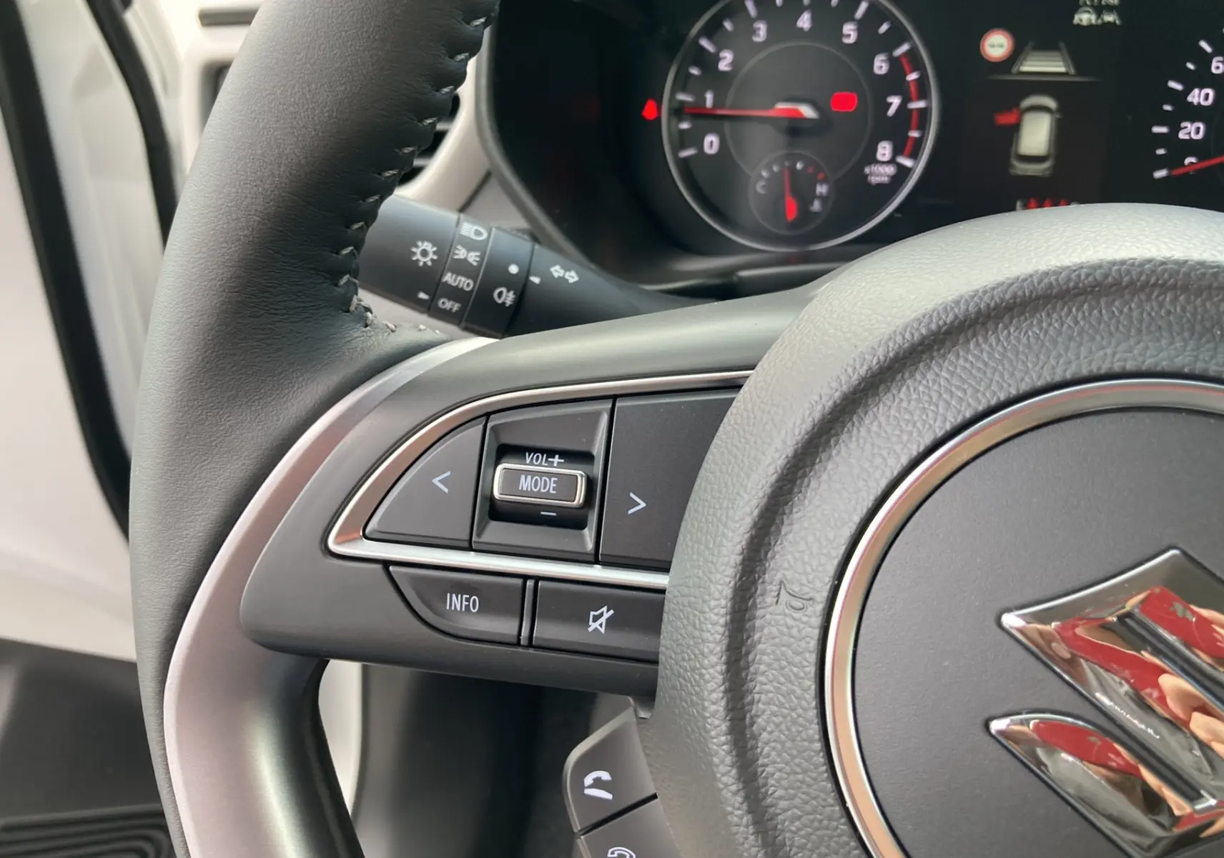 Gros plan sur le volant cuir multifonctions gris d’une Suzuki Swift blanche, avec commandes audio et tableau de bord en arrière-plan.