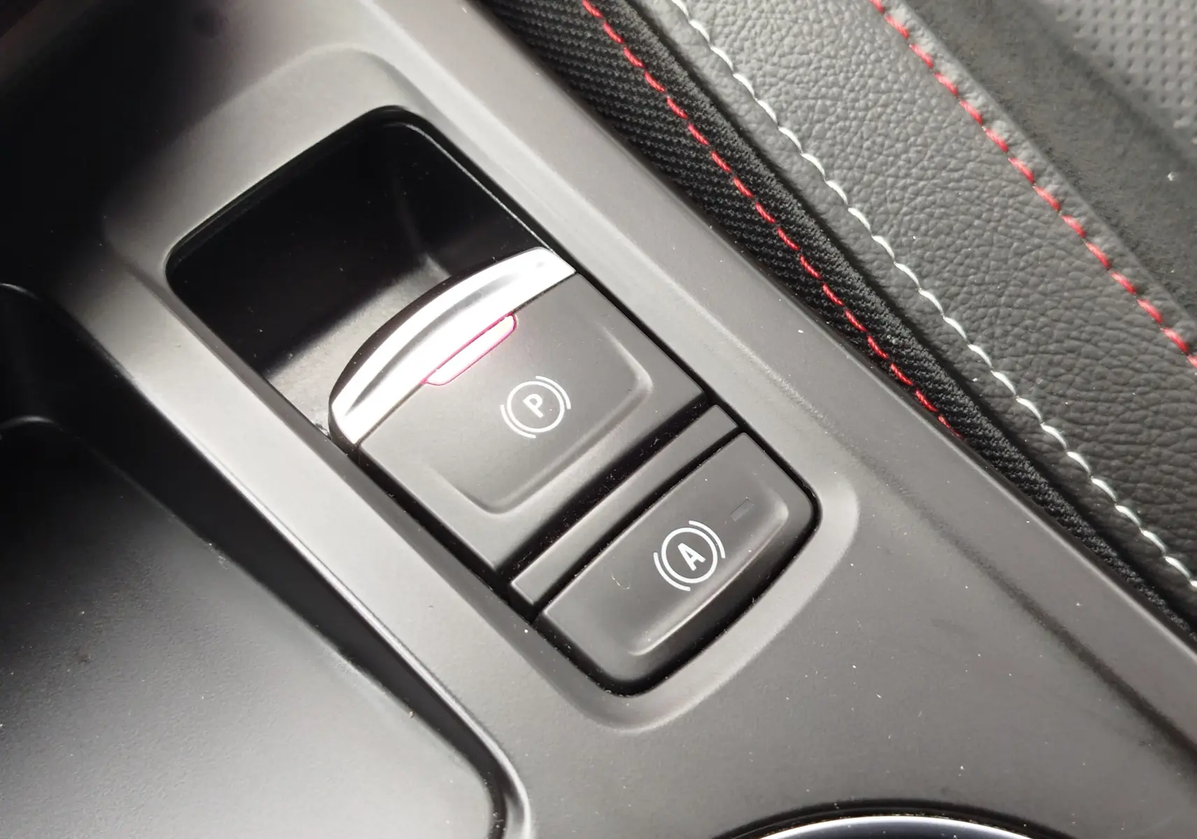 Gros plan sur les boutons de frein à main électronique et arrêt automatique du moteur, avec surpiqûres rouges sur cuir noir.