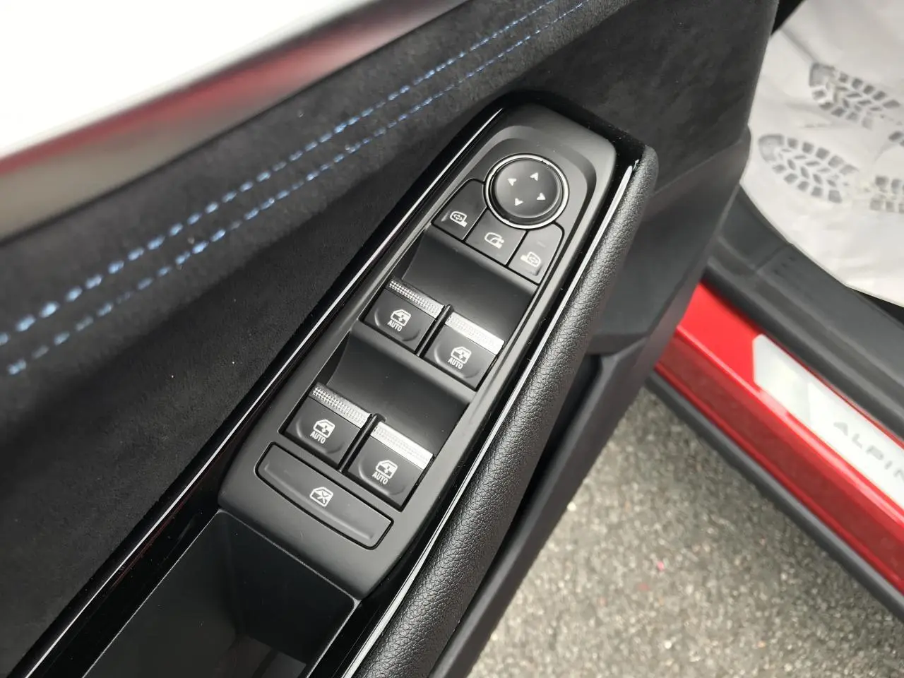 Gros plan sur les commandes de lève-vitres côté conducteur avec surpiqûres bleues et seuil de porte rouge Alpine visible.