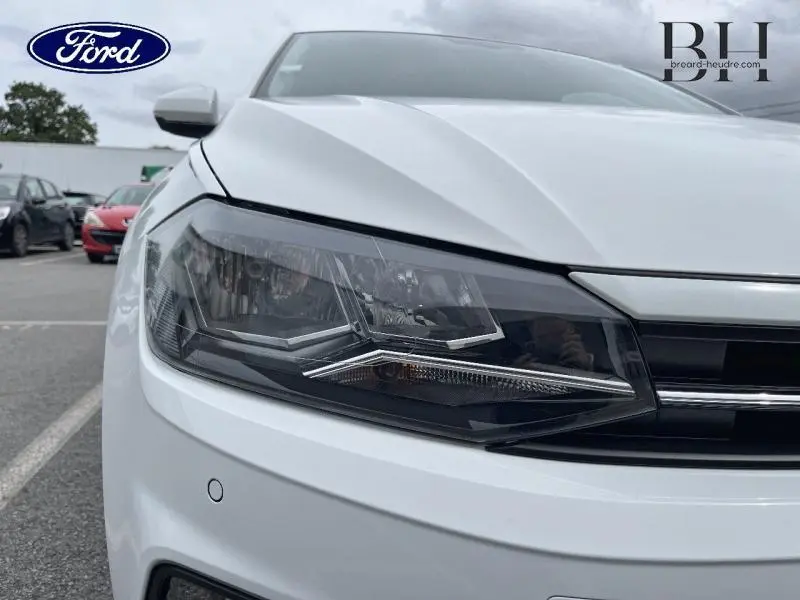 Gros plan sur le phare avant droit d'une Volkswagen Polo blanche Pure, soulignant la calandre chromée et le capteur de stationnement.