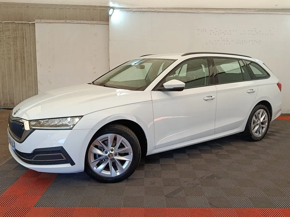 Skoda Octavia break blanc vue de profil droit en intérieur, avec jantes alliage et barres de toit noires.
