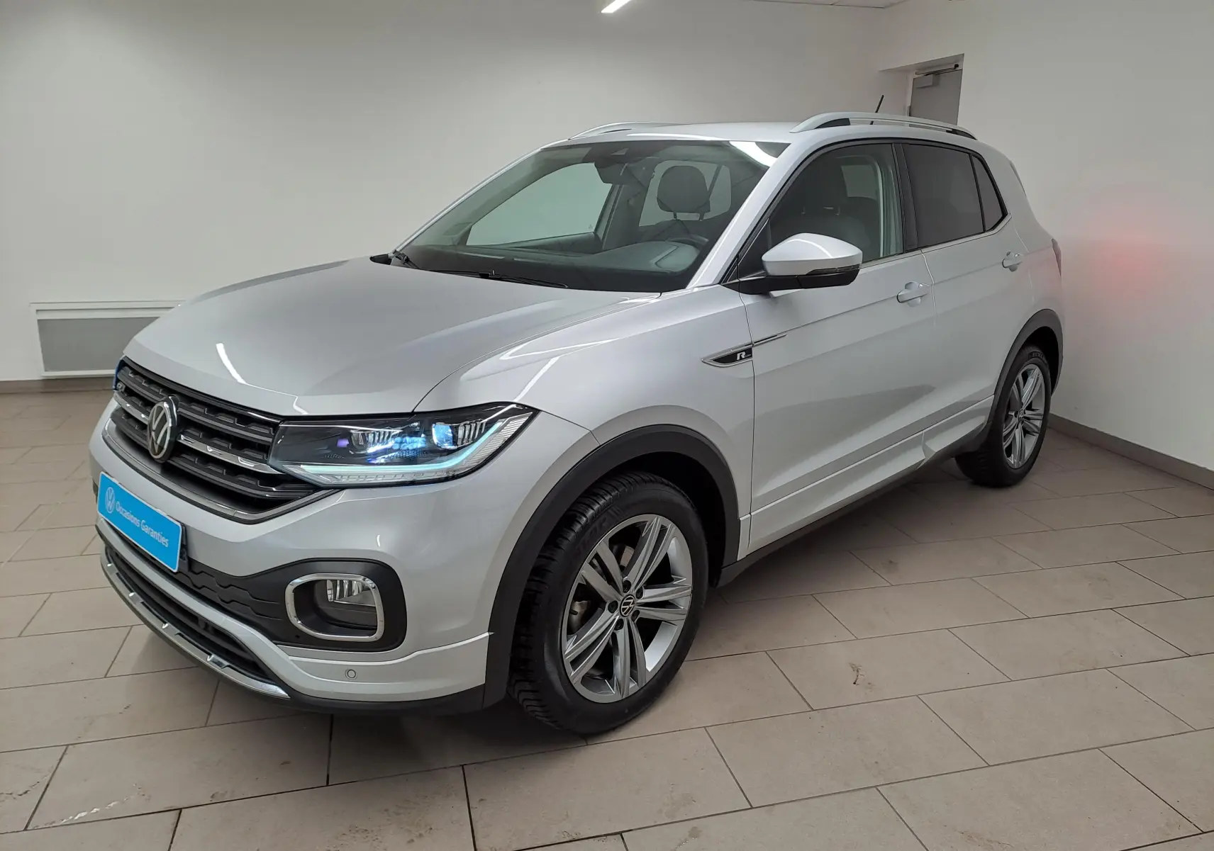 Volkswagen T-Cross 2022 argent reflet, vue 3/4 avant droit, avec badge R-Line et phares LED allumés.