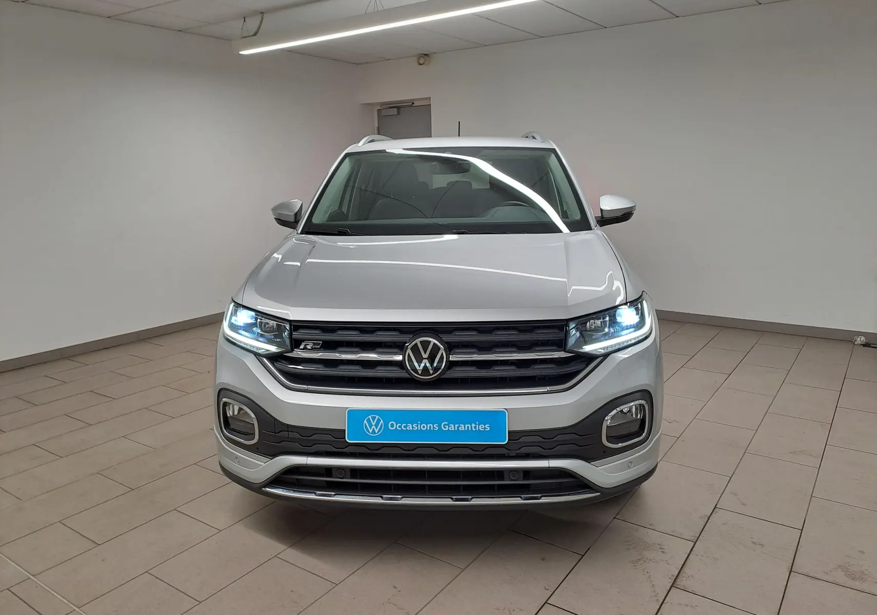 Volkswagen T-Cross 1.0 TSI R-Line 2022 en reflet d'argent, vue frontale avec phares allumés en intérieur.