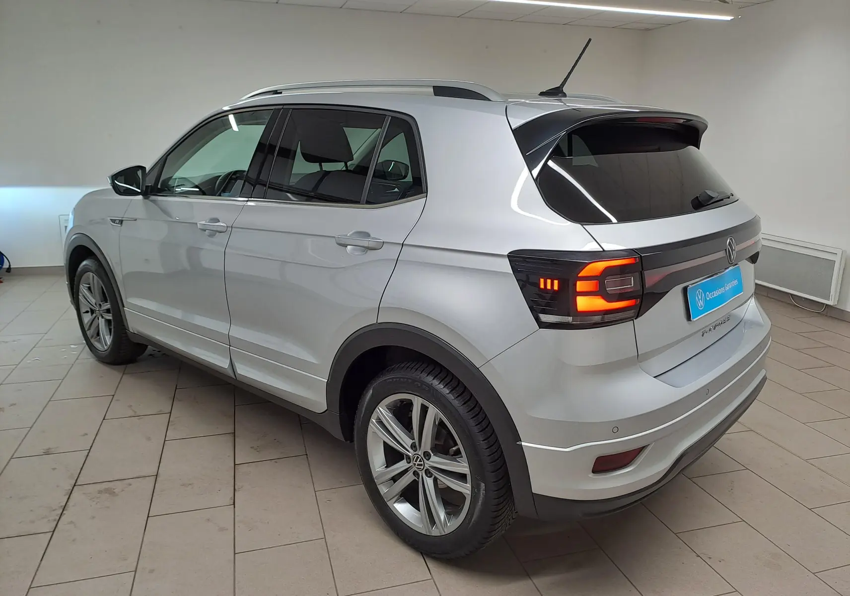 Vue 3/4 arrière côté gauche du Volkswagen T-Cross 1.0 TSI R-Line 2022 en argent avec feux arrière allumés.