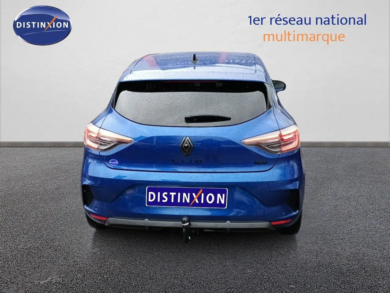 Vue arrière d'une Renault Clio 1.6 E-TECH hybride bleu Iron Metal avec attelage, sous une pluie légère.
