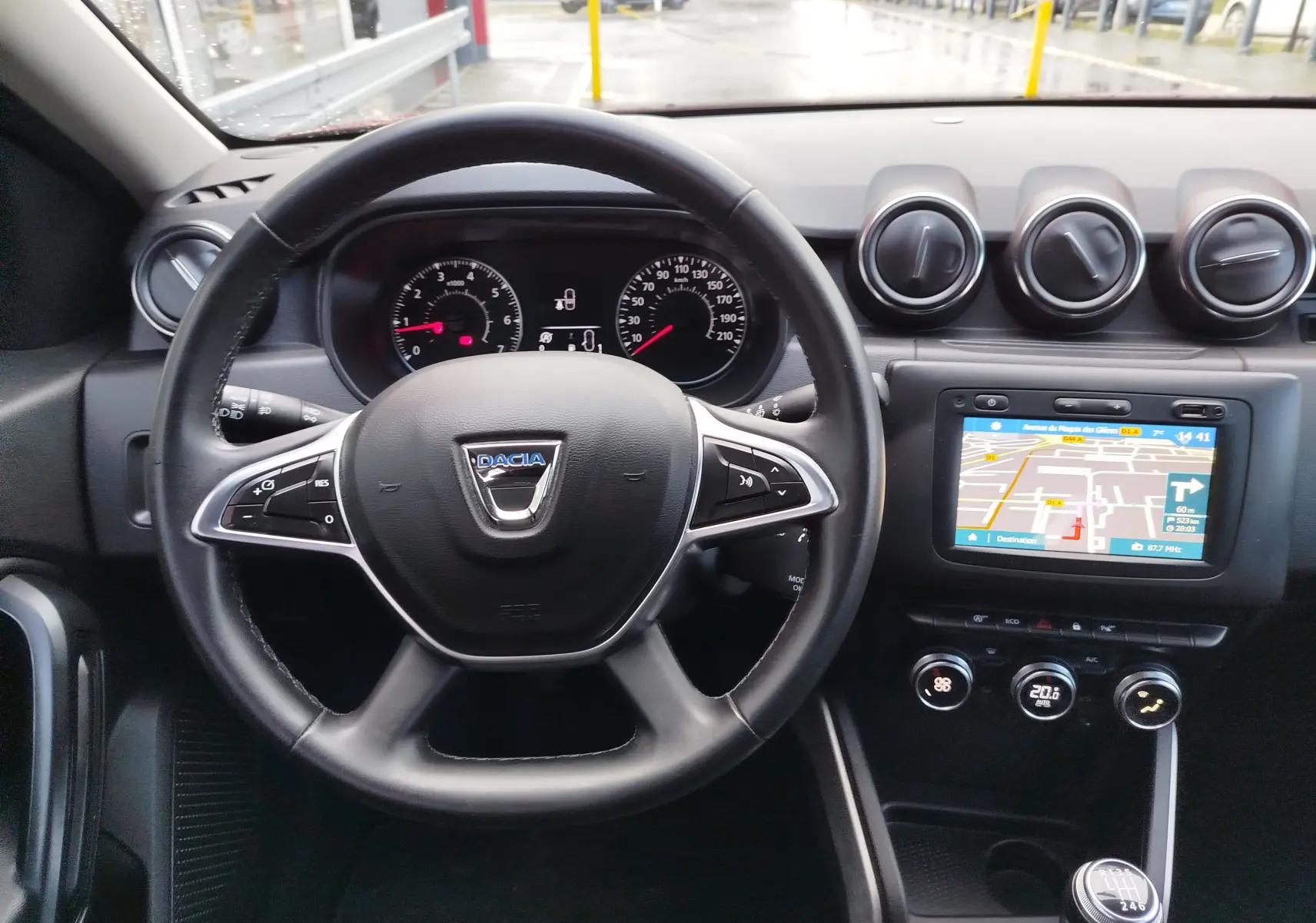 Vue intérieure centrée sur le volant Dacia, tableau de bord avec écran tactile et commandes de climatisation visibles.