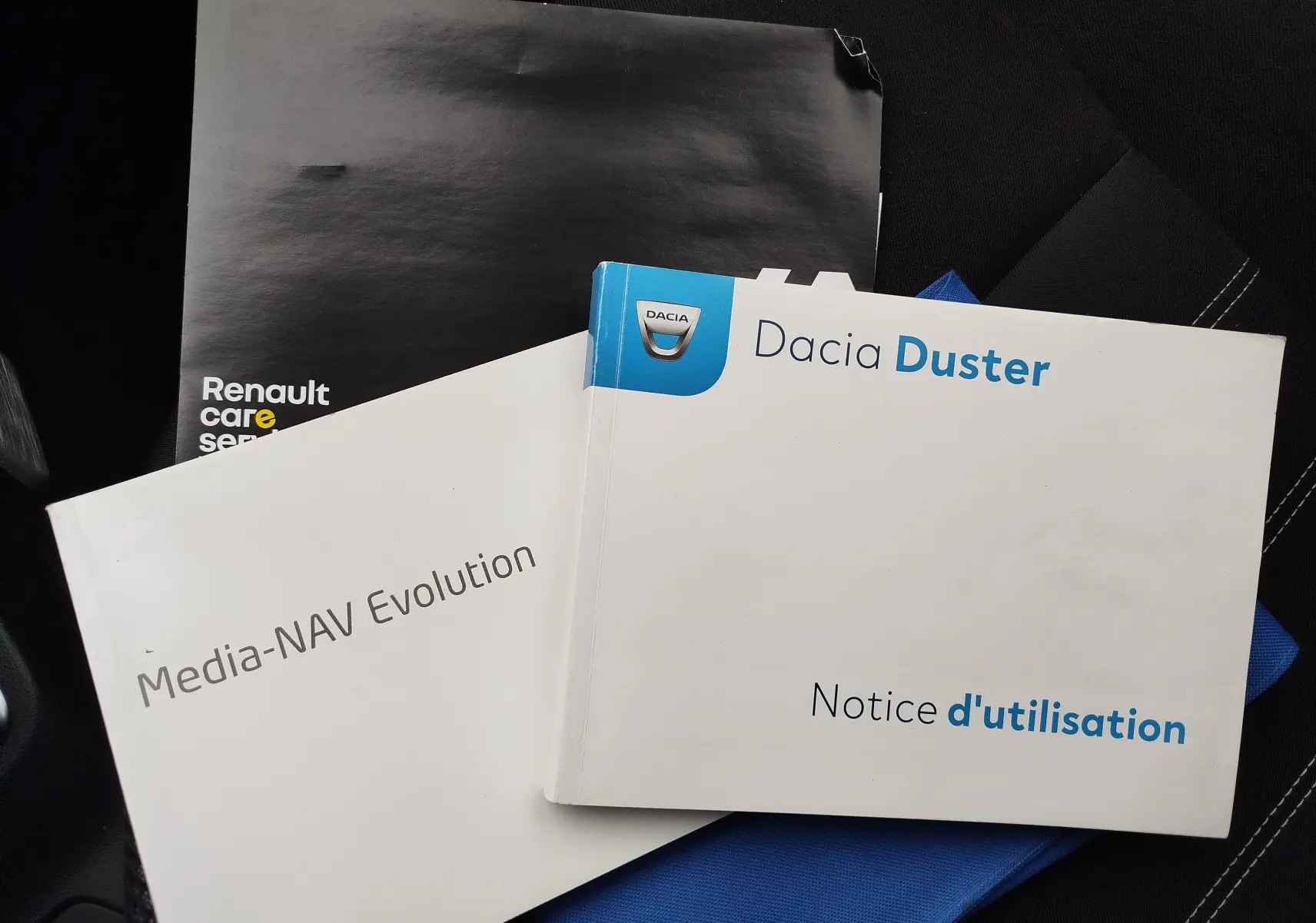 Image montrant trois manuels posés sur un siège de voiture, dont la notice d'utilisation du Dacia Duster en format paysage.