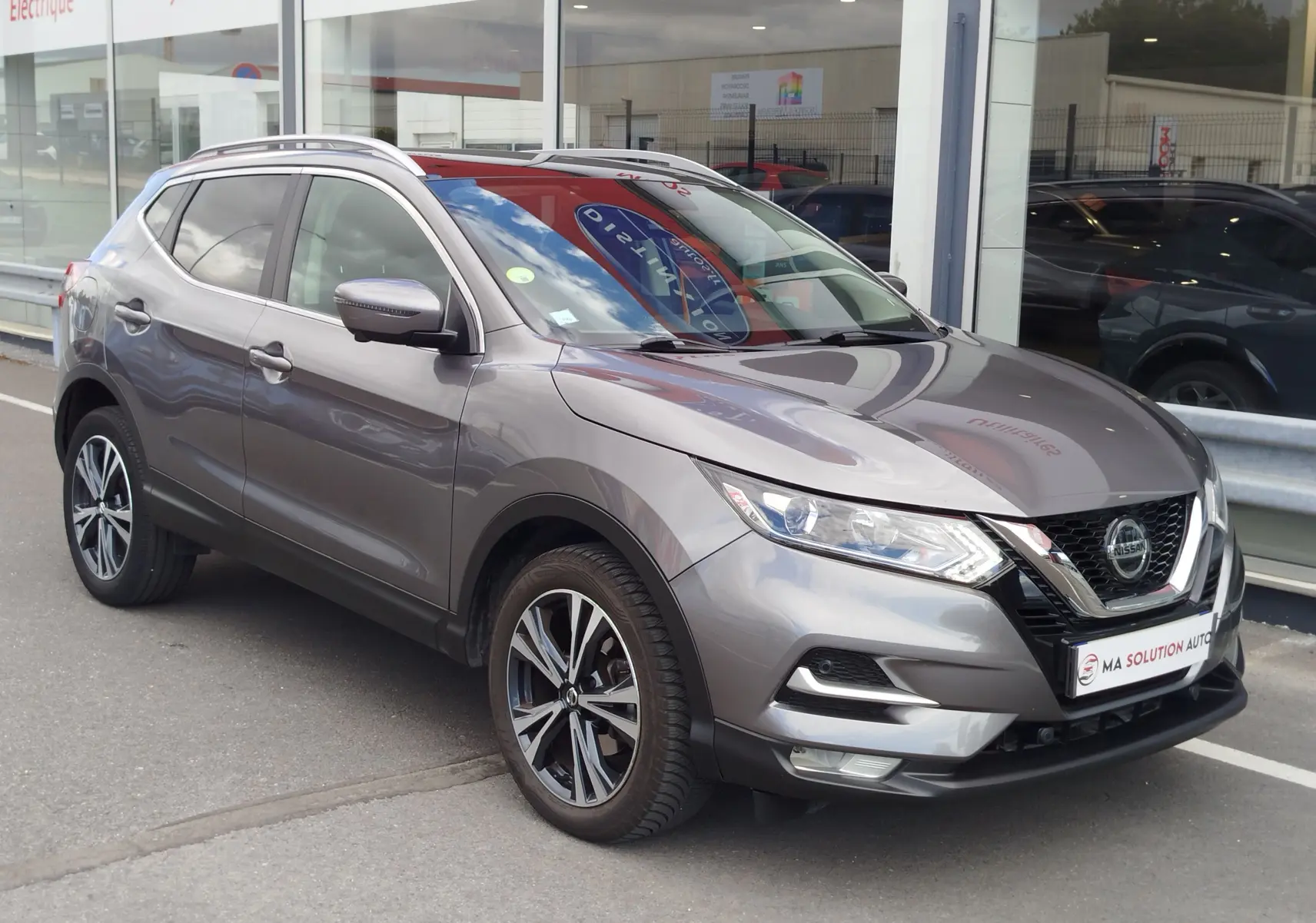 Nissan Qashqai gris Squale en 3/4 avant droit, avec jantes alliage et calandre chromée visible devant un showroom.