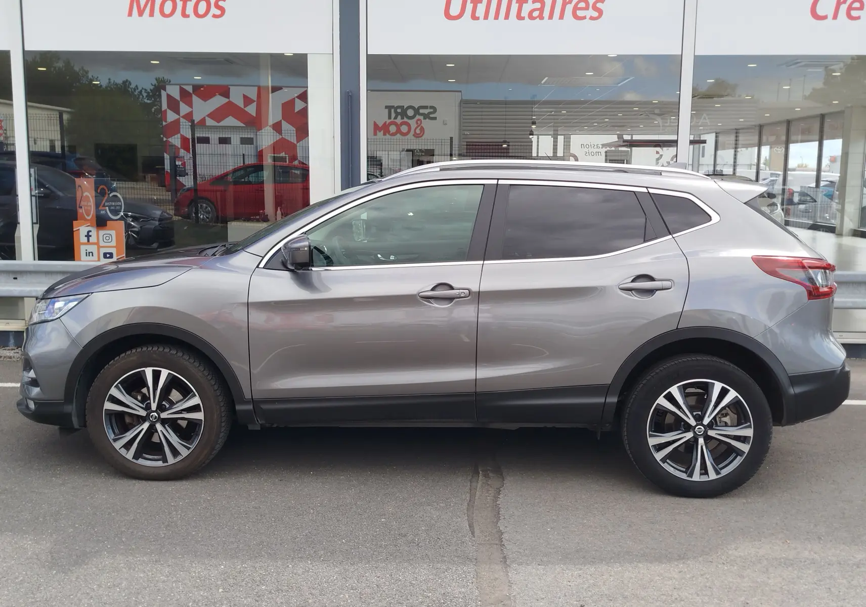 Vue de profil côté gauche d'un Nissan Qashqai gris Squale avec jantes alliage et vitres teintées, stationné devant un showroom.
