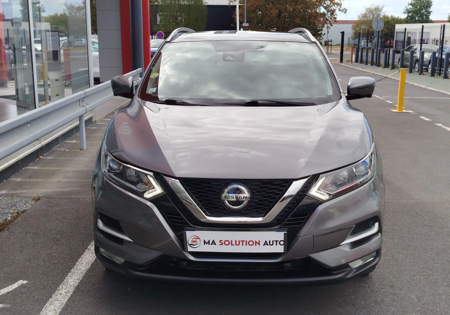 Vue avant d'un Nissan Qashqai gris Squale 2021 avec feux LED allumés et calandre chromée, stationné devant un bâtiment.