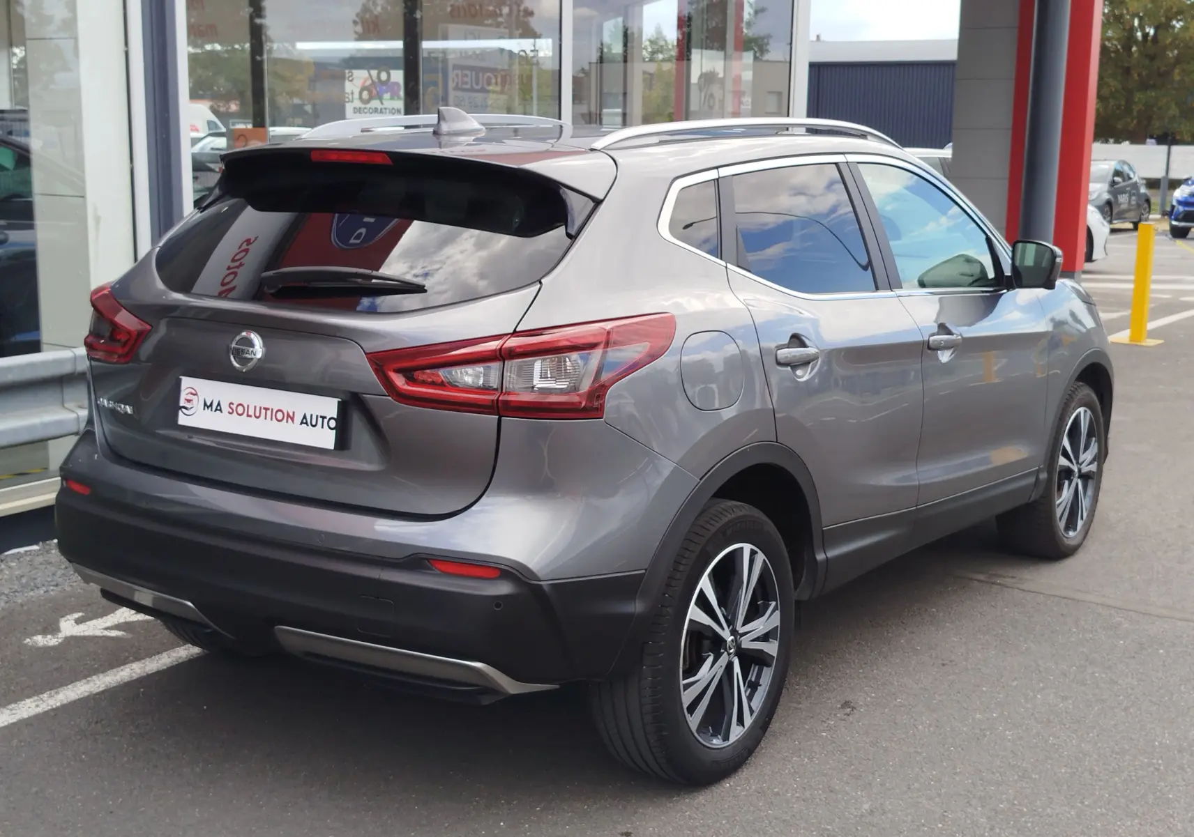 Nissan Qashqai gris Squale vu en 3/4 arrière droit, avec jantes alliage et vitres teintées, stationné devant un showroom.