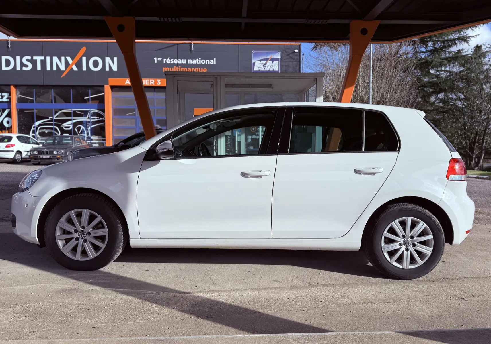 Profil côté gauche d'une Volkswagen Golf blanche 5 portes de 2012, stationnée devant un bâtiment commercial.