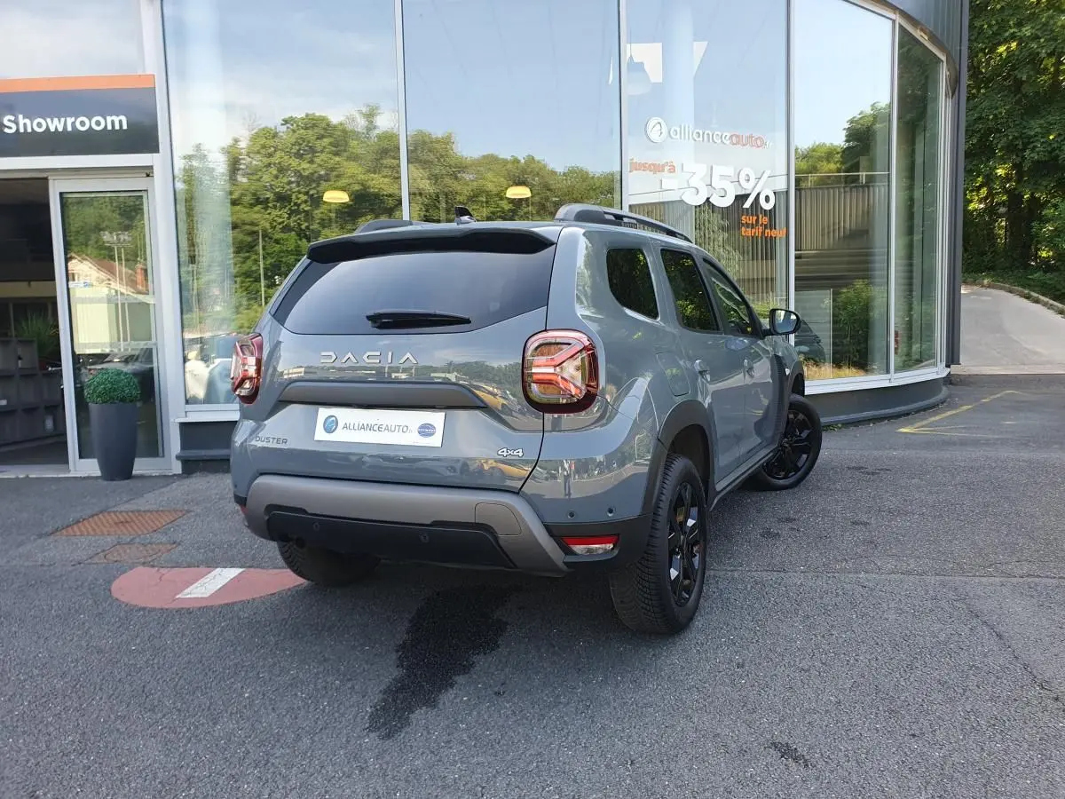 Vue 3/4 arrière droite d'un Dacia Duster gris urbain 2023 avec jantes noires et vitres surteintées, stationné devant un showroom.