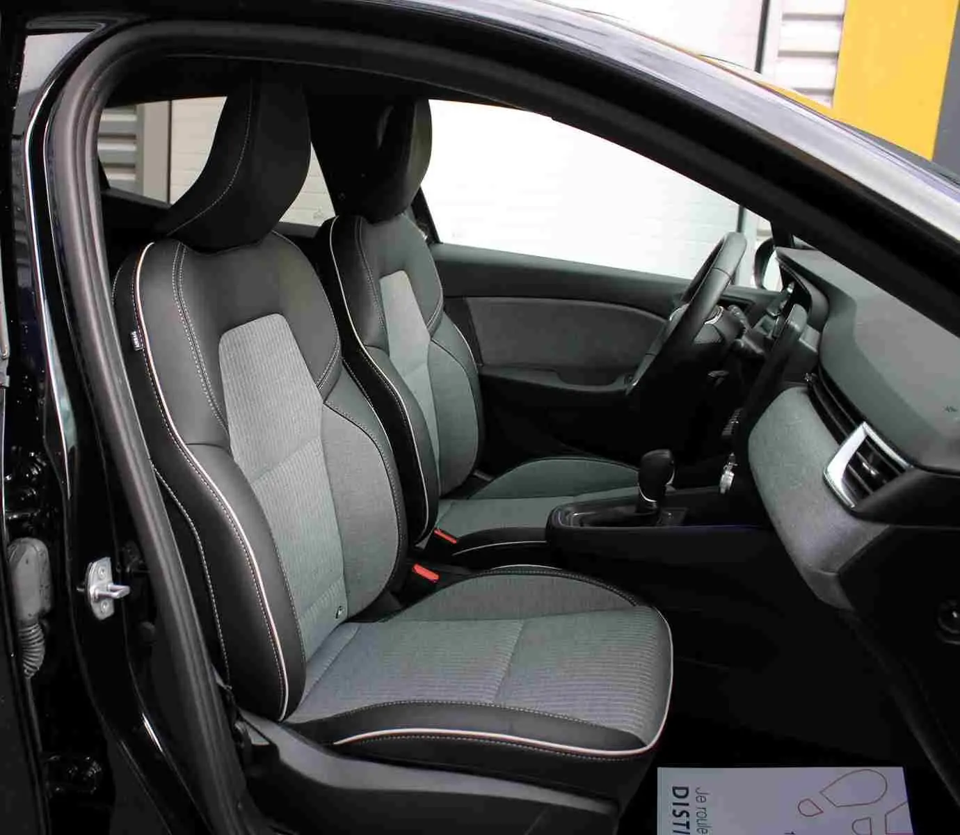 Vue intérieure côté conducteur de la Renault Clio noire étoile métal, sièges tissu gris noir avec surpiqûres blanches.