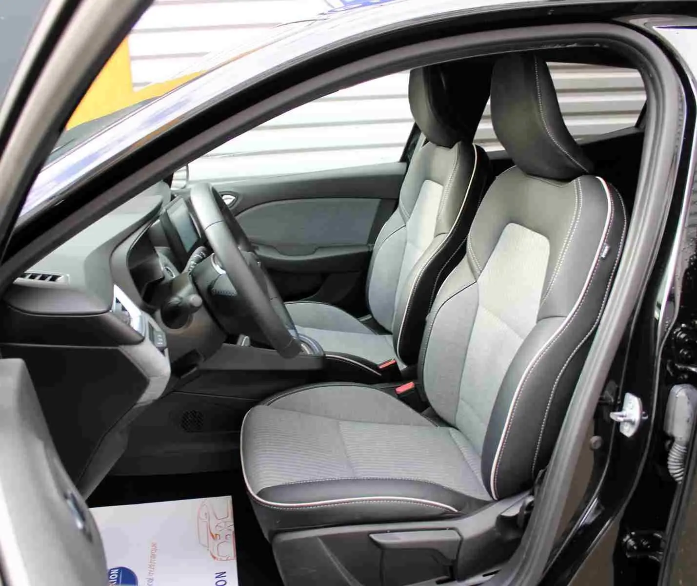 Vue intérieure côté conducteur de la Renault Clio 2025 noire étoile métal, sièges tissu gris clair avec surpiqûres blanches.