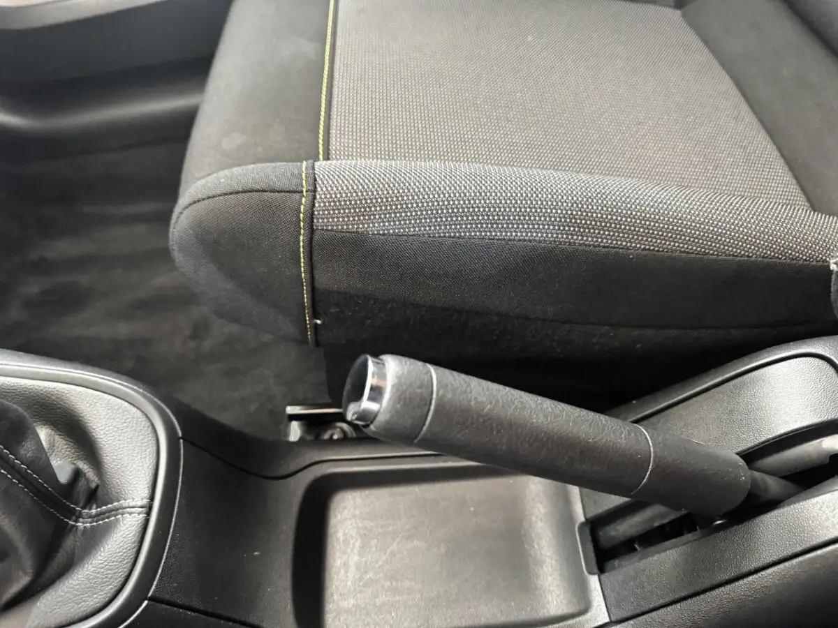 Gros plan sur le frein à main et la console centrale noire, avec siège tissu gris foncé à surpiqûres jaunes visible côté droit.