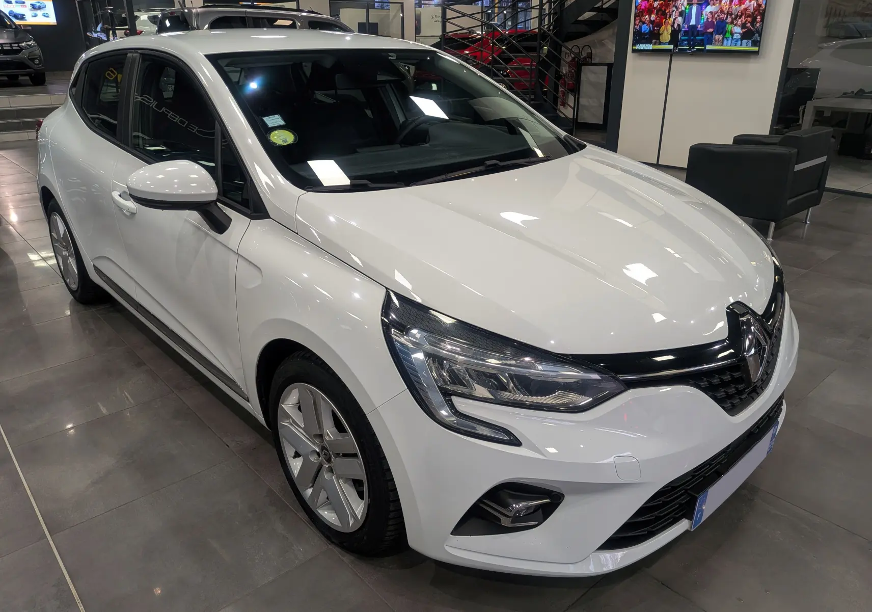 Renault Clio V Blue dCi 115 Business blanc glacier vue 3/4 avant droit en showroom avec phares LED distinctifs.