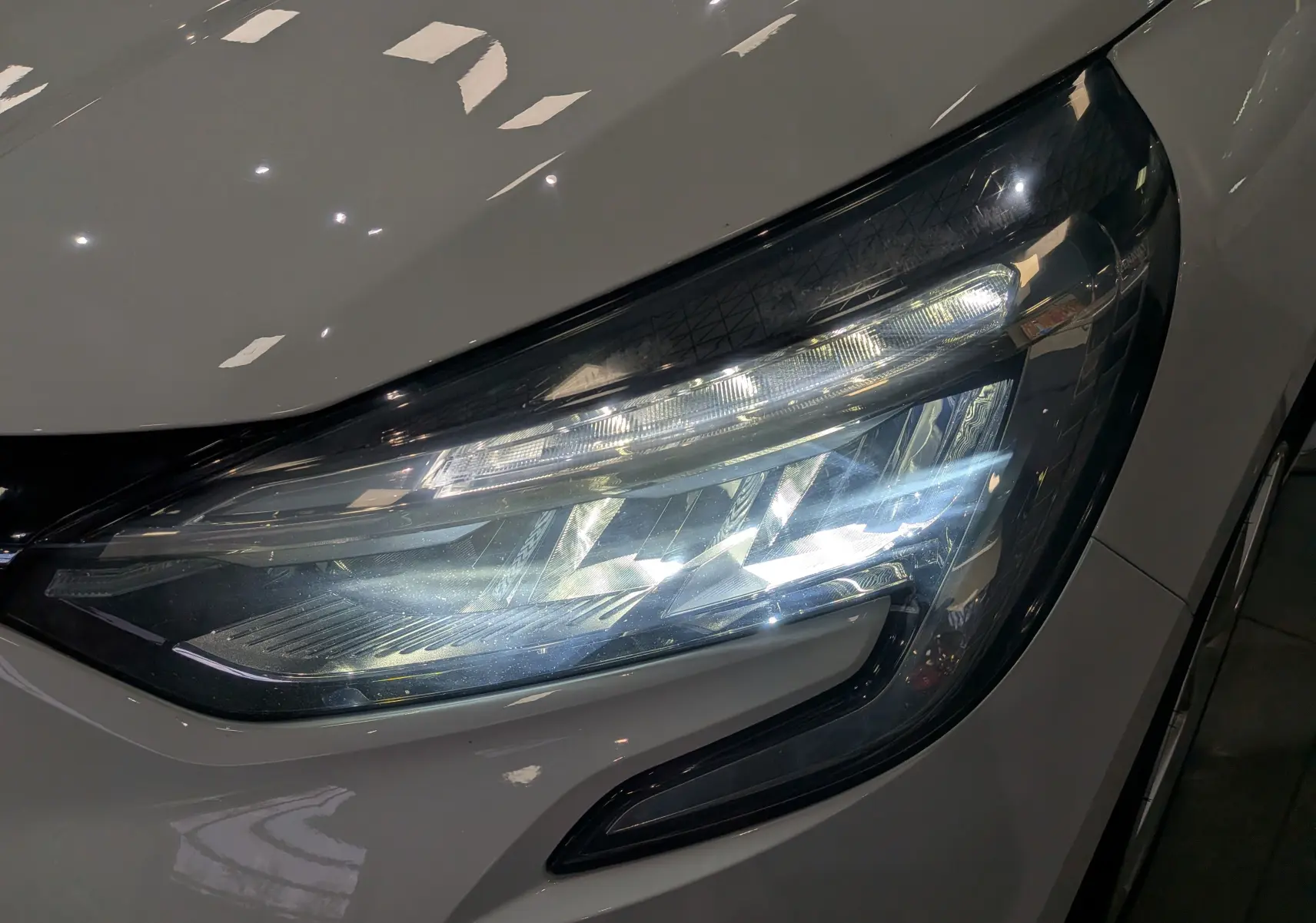 Gros plan sur le phare avant droit allumé d'une Renault Clio V Blanc Glacier, soulignant son design LED moderne.