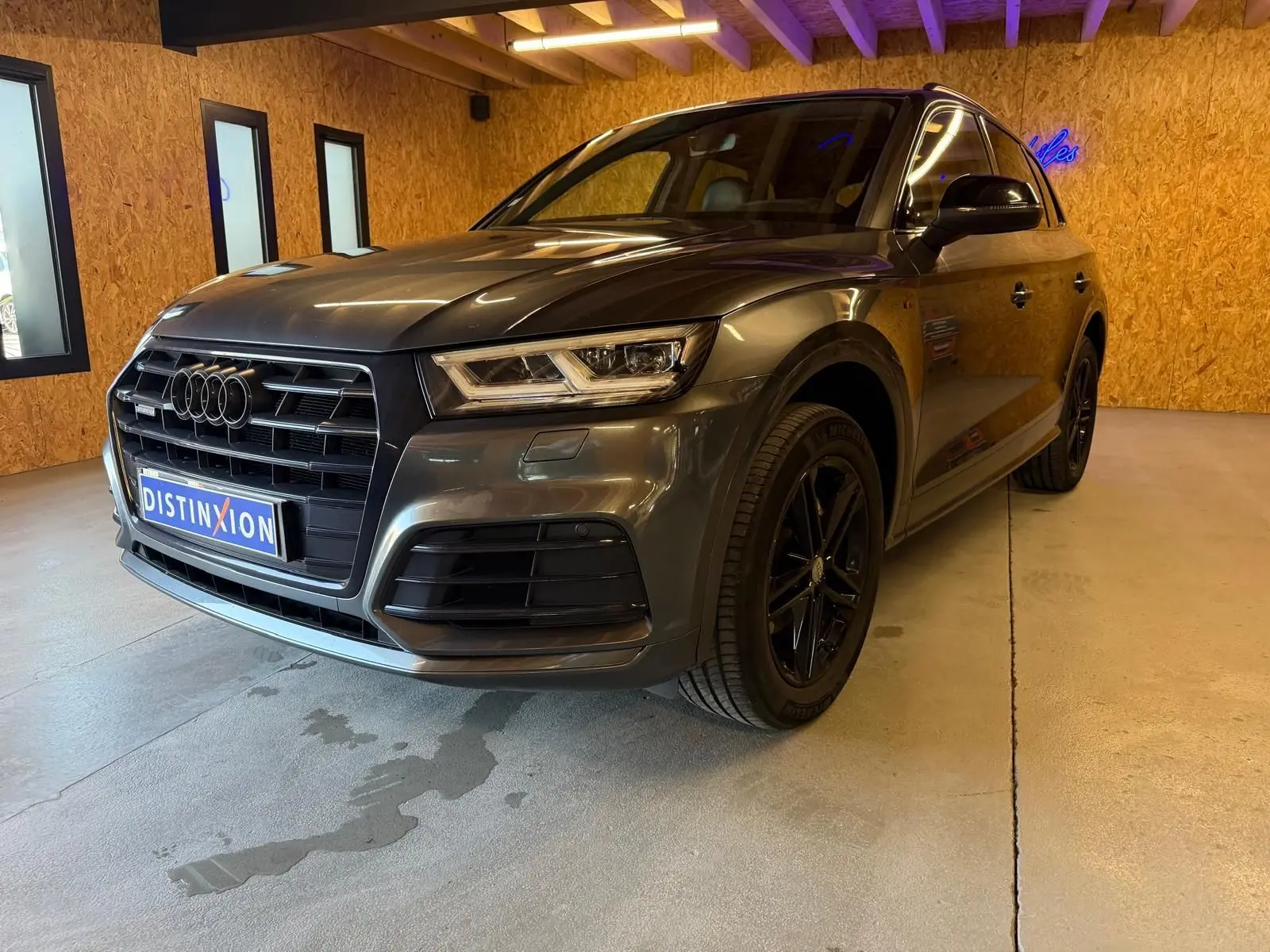 Audi Q5 Sportback gris en vue 3/4 avant droit, avec jantes noires et calandre imposante dans un intérieur en bois.