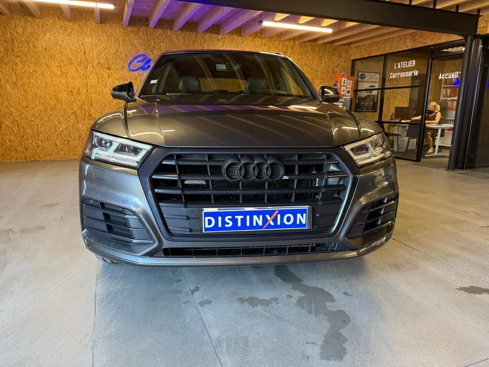 Vue avant d'un Audi Q5 Sportback gris 2019 avec calandre noire et plaques Distinxion en intérieur d'atelier.