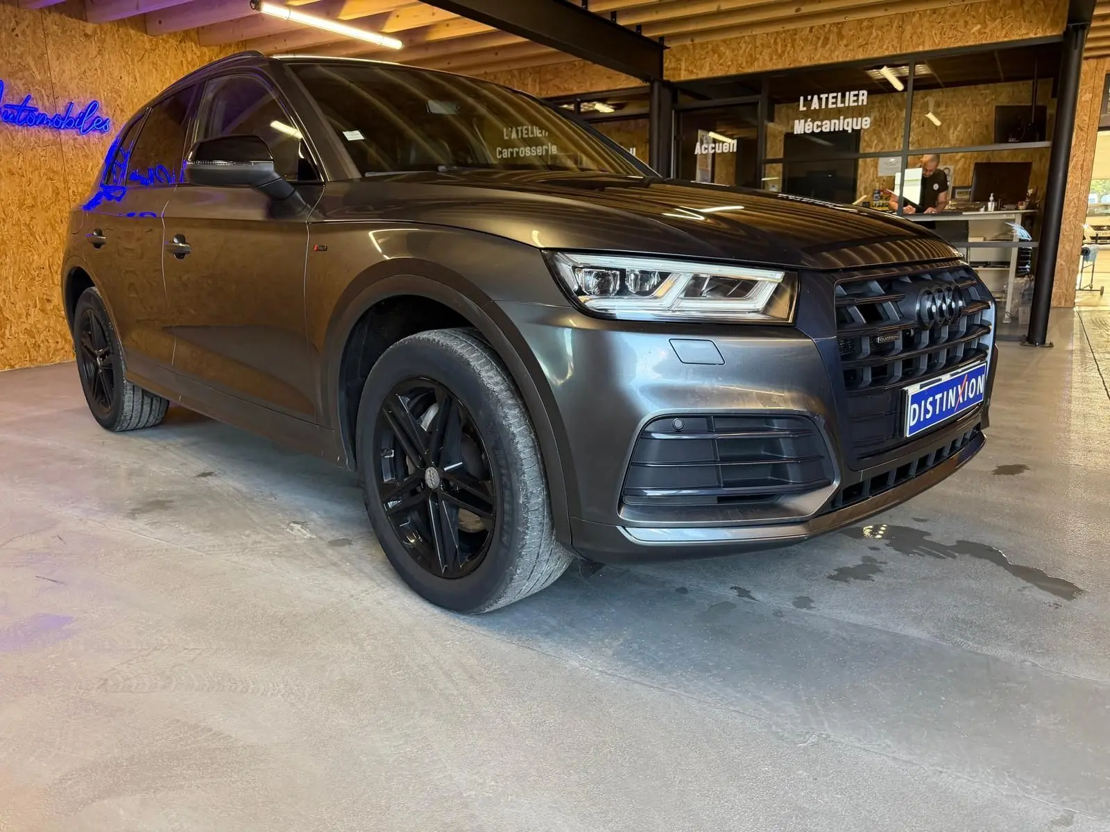 Audi Q5 Sportback gris en 3/4 avant droit, avec jantes noires et calandre marquée S-Line dans un garage.