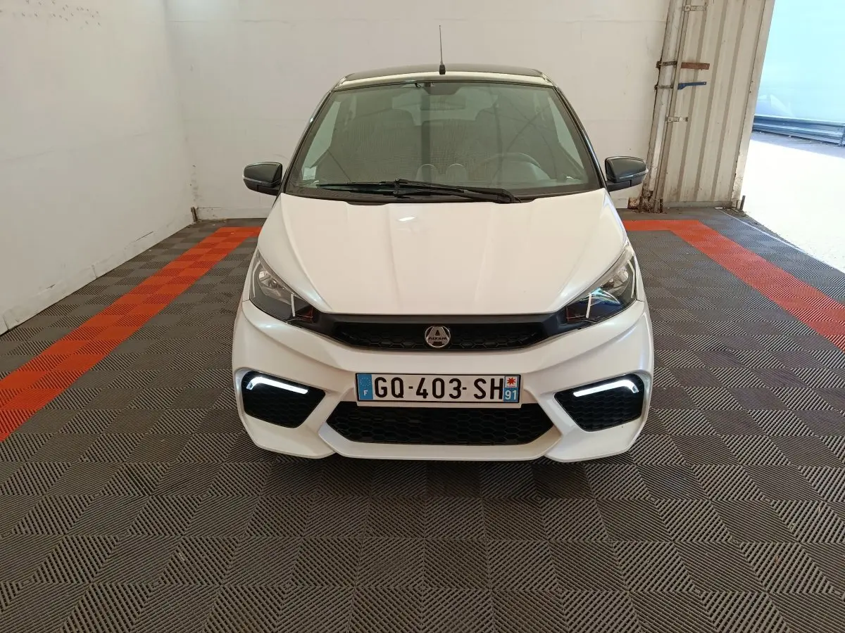 Vue de face d'une Aixam Ambition 0.5 City Coupe GTI blanche avec feux LED diurnes allumés dans un garage.