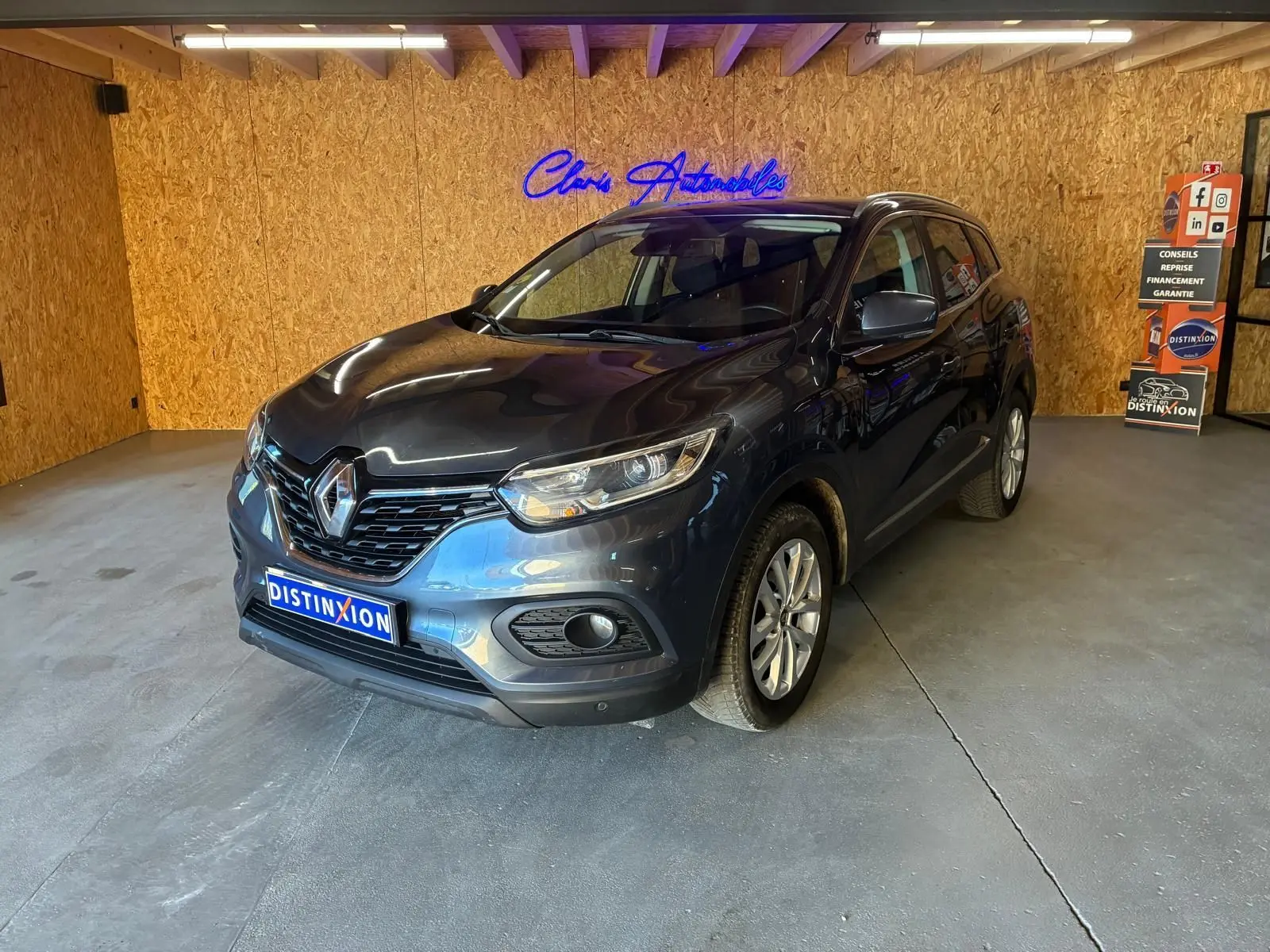 Renault Kadjar gris 2019 en 3/4 avant droit dans un garage avec mur en bois et enseigne lumineuse bleue.