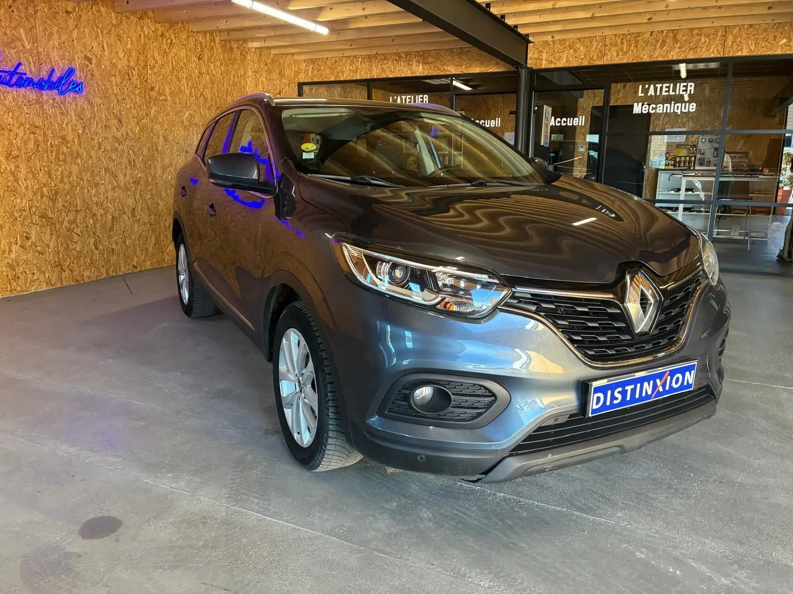 Renault Kadjar gris 3/4 avant droit, SUV compact avec calandre noire et phares LED, stationné en intérieur showroom.