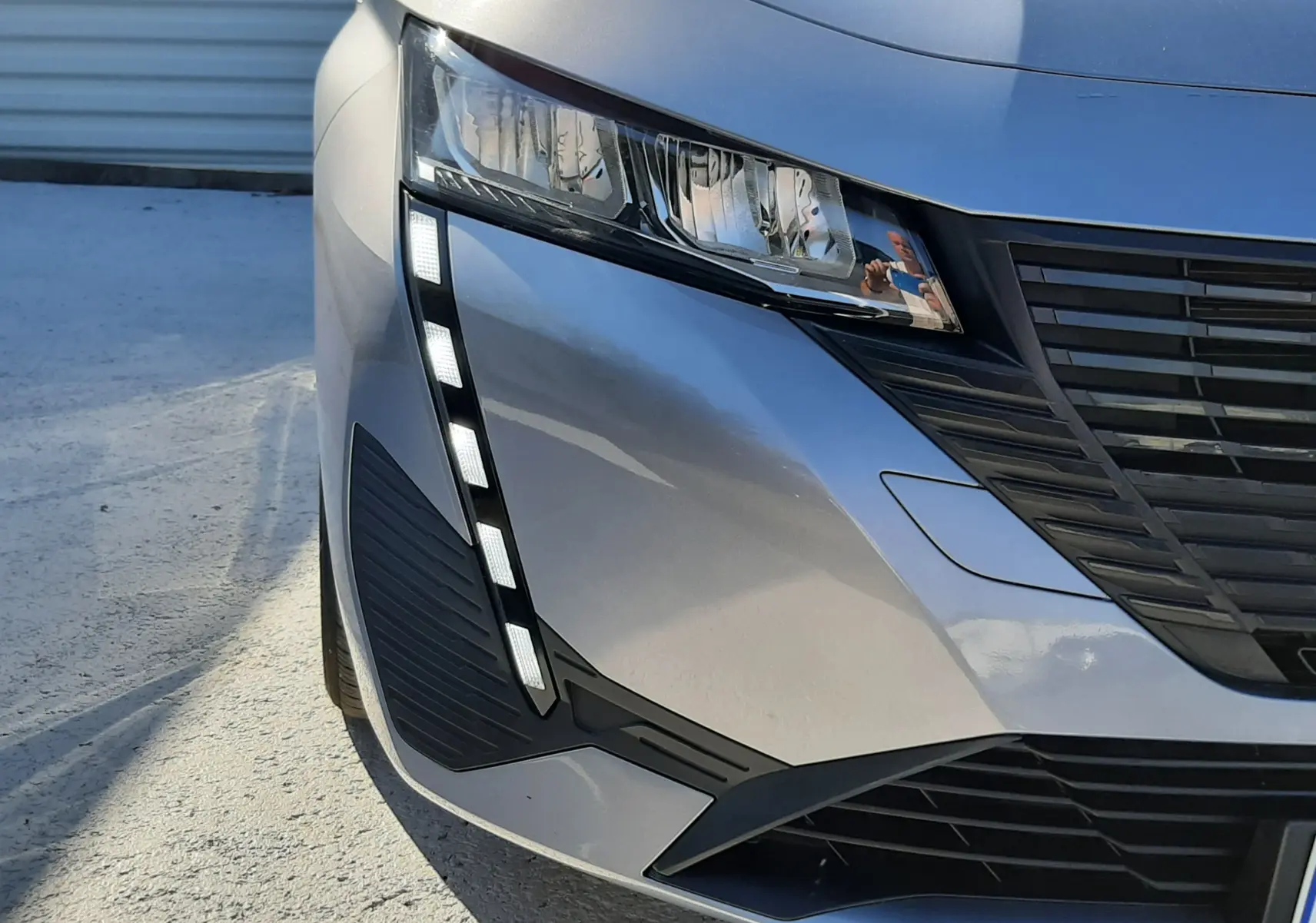 Gros plan sur l'avant droit gris Artense du Peugeot 308 III Hybrid 145ch, mettant en valeur le phare et la calandre noire.