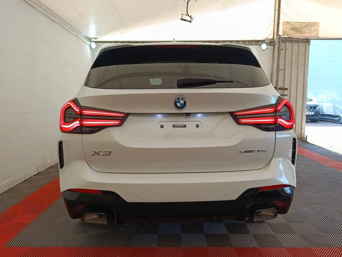 Vue arrière d’un BMW X3 xDrive30e blanc avec feux LED allumés et double sortie d’échappement dans un garage.