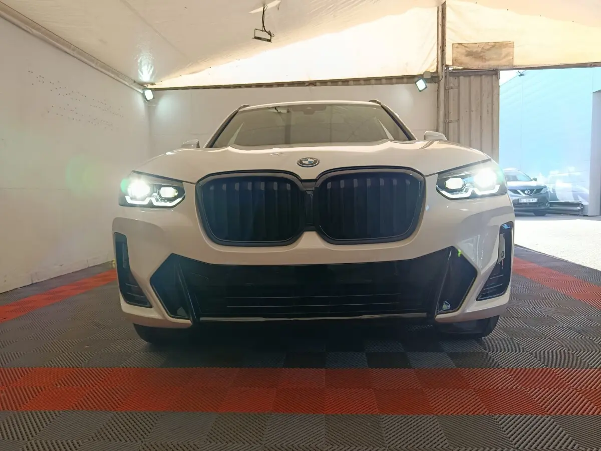 Vue frontale d'un BMW X3 blanc avec phares allumés et calandre noire dans un garage couvert.
