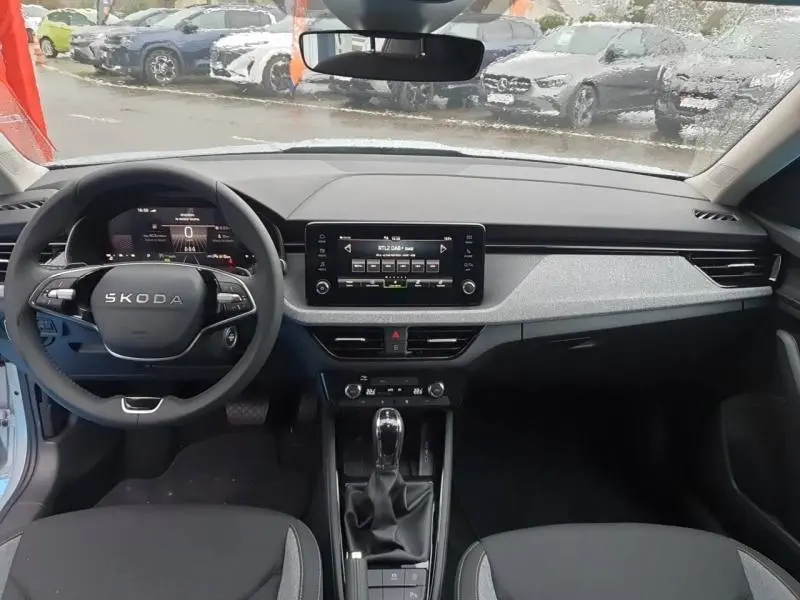 Vue intérieure avant du Skoda Kamiq 2025, tableau de bord gris clair avec écran tactile et volant multifonction noir.