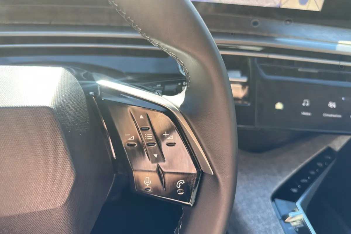 Gros plan sur les commandes du volant cuir multifonctions du Peugeot 5008 Hybrid 145 gris Artense, tableau de bord numérique en arrière-plan.