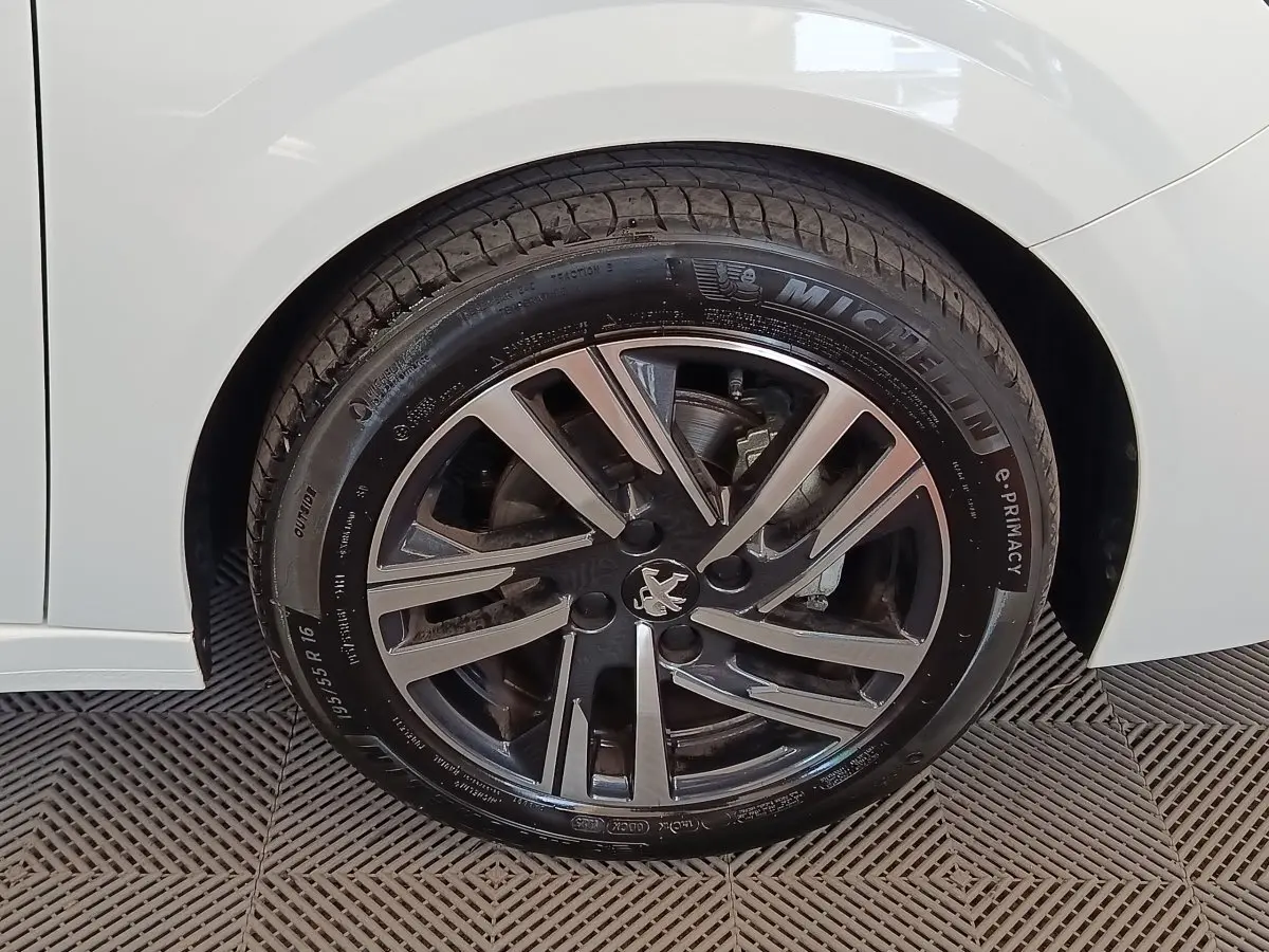 Gros plan sur la roue avant gauche d'une Peugeot 208 blanche, jante alliage au design moderne avec logo lion central.