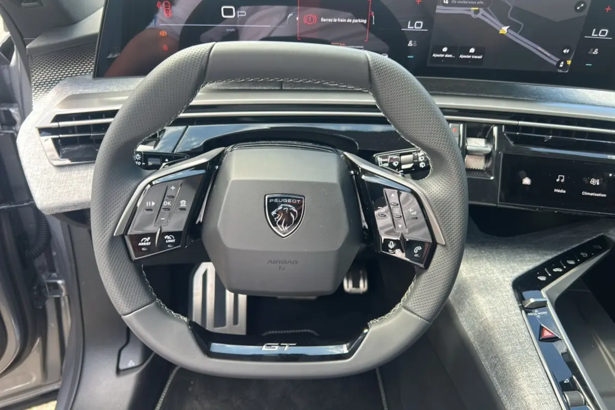 Vue rapprochée du volant cuir multifonctions GT du Peugeot 5008 Hybrid 145 gris Artense avec tableau de bord numérique.