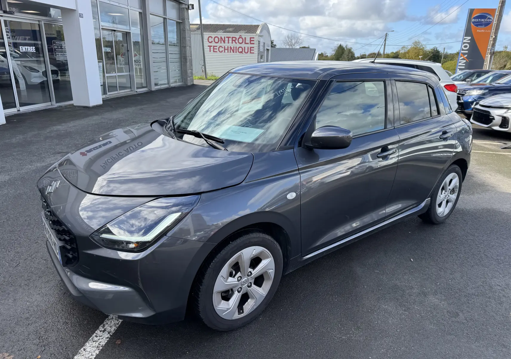 Suzuki Swift 1.2 Hybrid Privilege gris vue 3/4 avant droit, avec jantes alliage et calandre noire brillante.