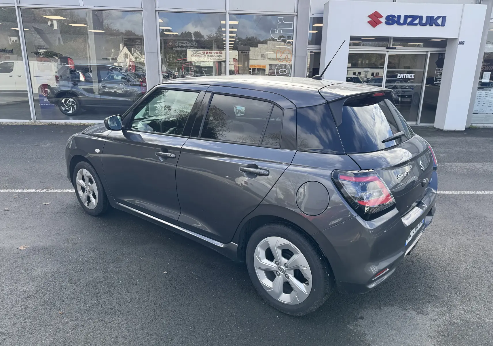 Suzuki Swift 1.2 Hybrid Privilege gris vue 3/4 arrière droit, stationnée devant un concessionnaire Suzuki.